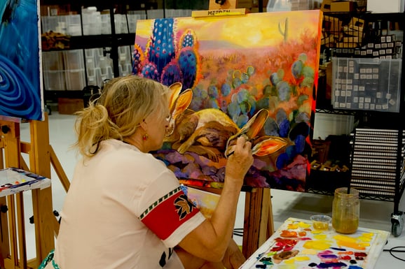 Woman Mary Ambrose painting desert scene of blue cactus and brown rabbit