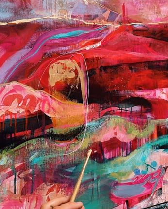 hand holding a paint brush in front of a colorful abstract canvas red blue gold