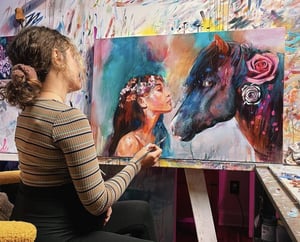 girls in tan shirt painting sitting in frint of painting of a brown horse and girl with flower crwon on 