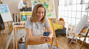 Woman artist in gray shirt sitting in art studio on phone smiling 