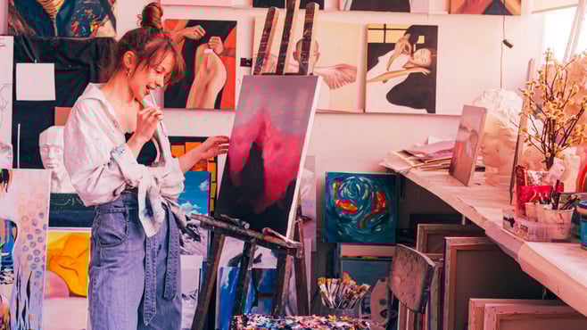 Woman standing in front of painting on easel blue pink cream