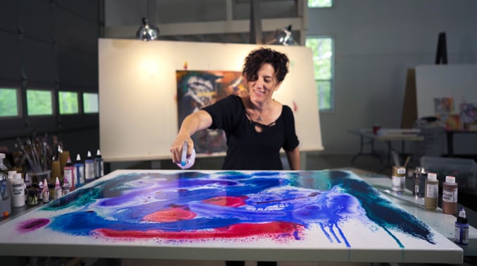 Artist smiling while using a spray bottle to blend vibrant blue, red, and purple inks on a large canvas in a well-lit art studio, surrounded by paint bottles and brushes