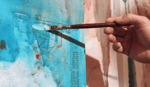Hand holding a paintbrush with paint on a teal canvas