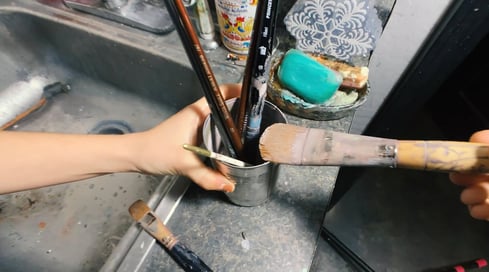 Hands holding dirty paint brushes near sink