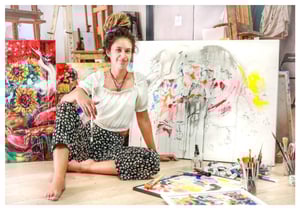 Woman sitting in front of paining holding paint brush 