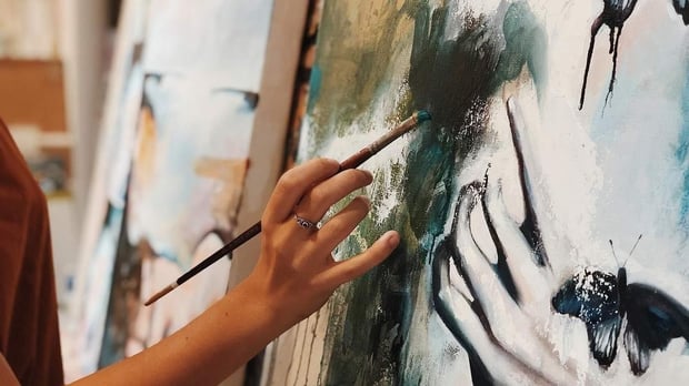 Hand holding paint brush to large canvas canvas has earth tone colors and painting of hand and butterfly