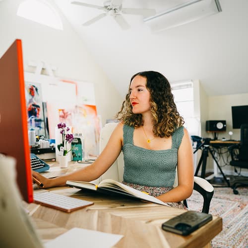 woman in gray sitting at desk with orange computer and book