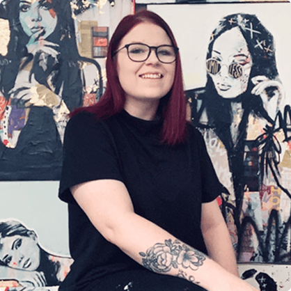 Tanya Aubut, artist in black shirt sitting in front of paintings of women