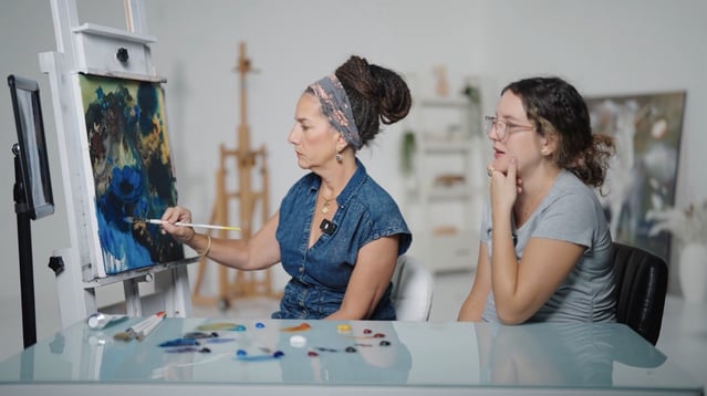 Woman in blue teaching woman in grey at art easel
