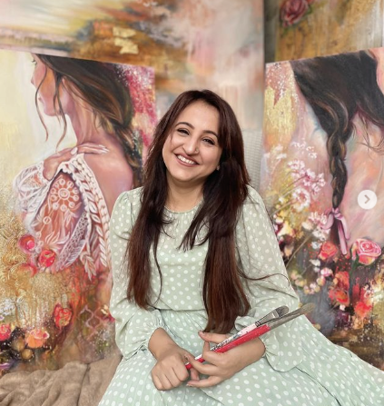 Ritika Aurora sitting in fron of pink tan brown yellow painitngs holding paint brushes