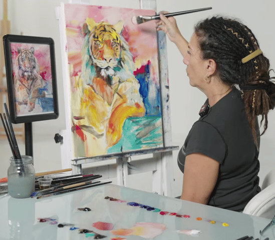 woman painitng yellow tiger on canvas with blue green pink background