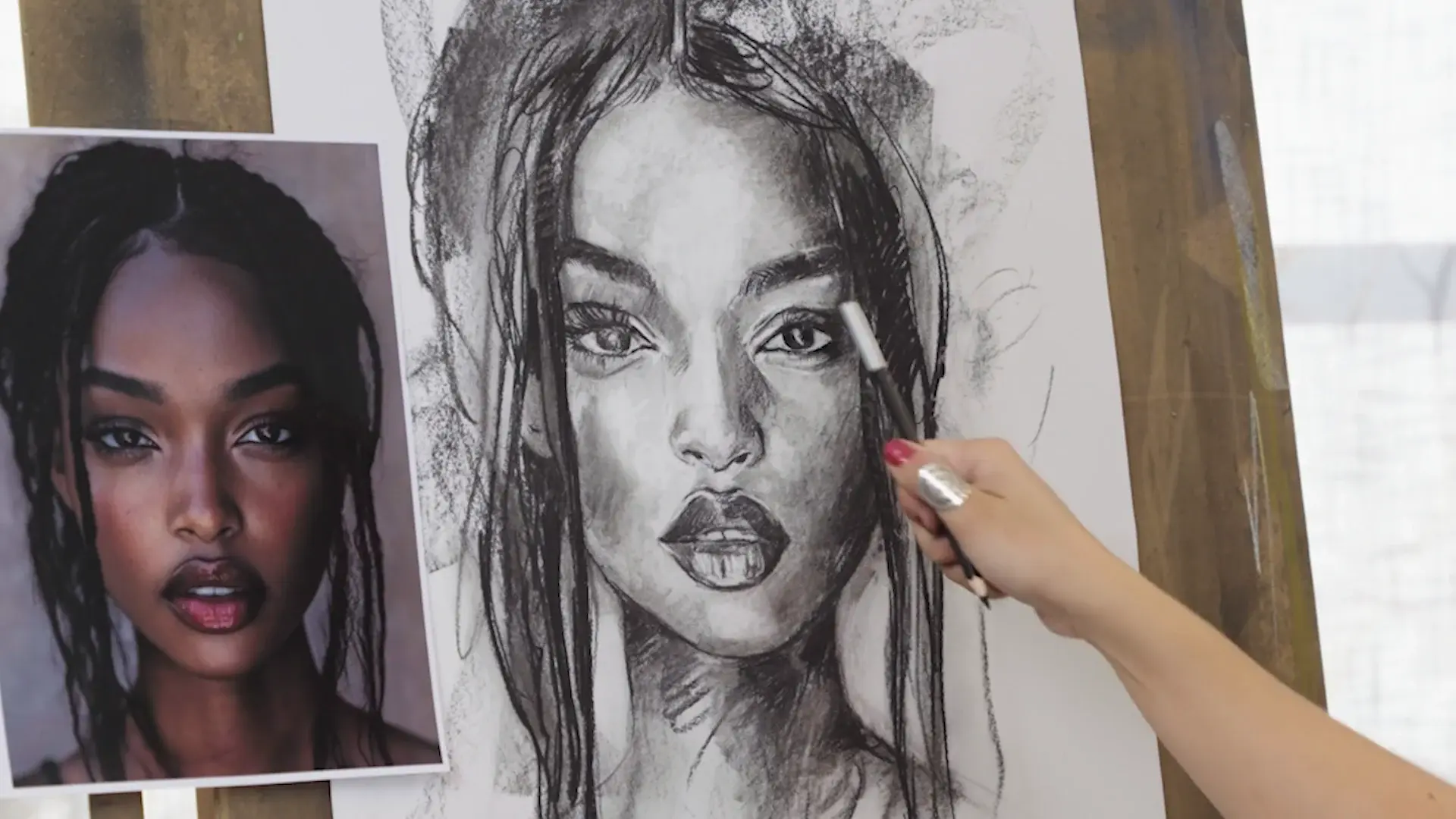 The nearly finished charcoal portrait beside its reference photo, a white charcoal pencil in hand