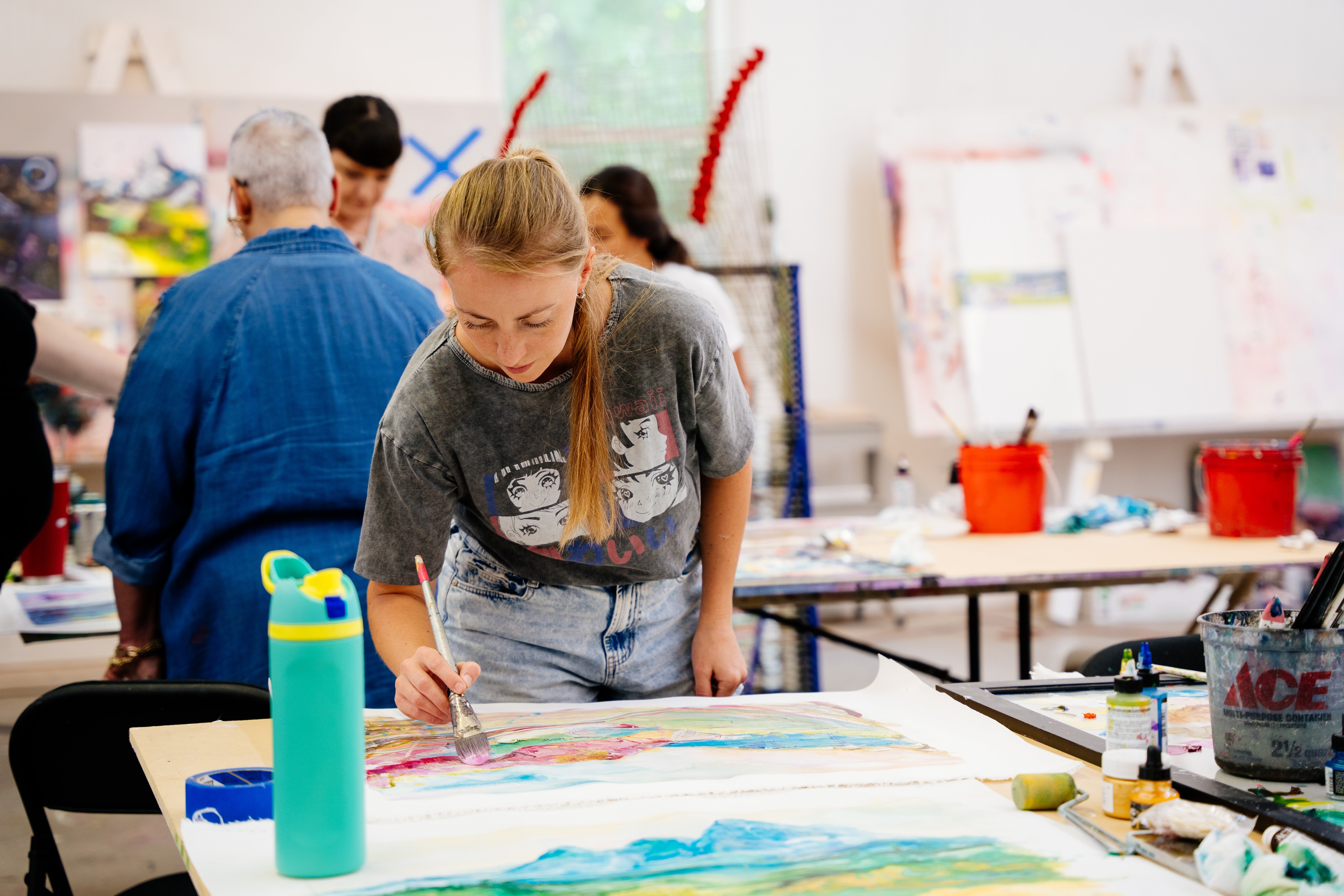 Woman in grey shirt bent over table painting landscape