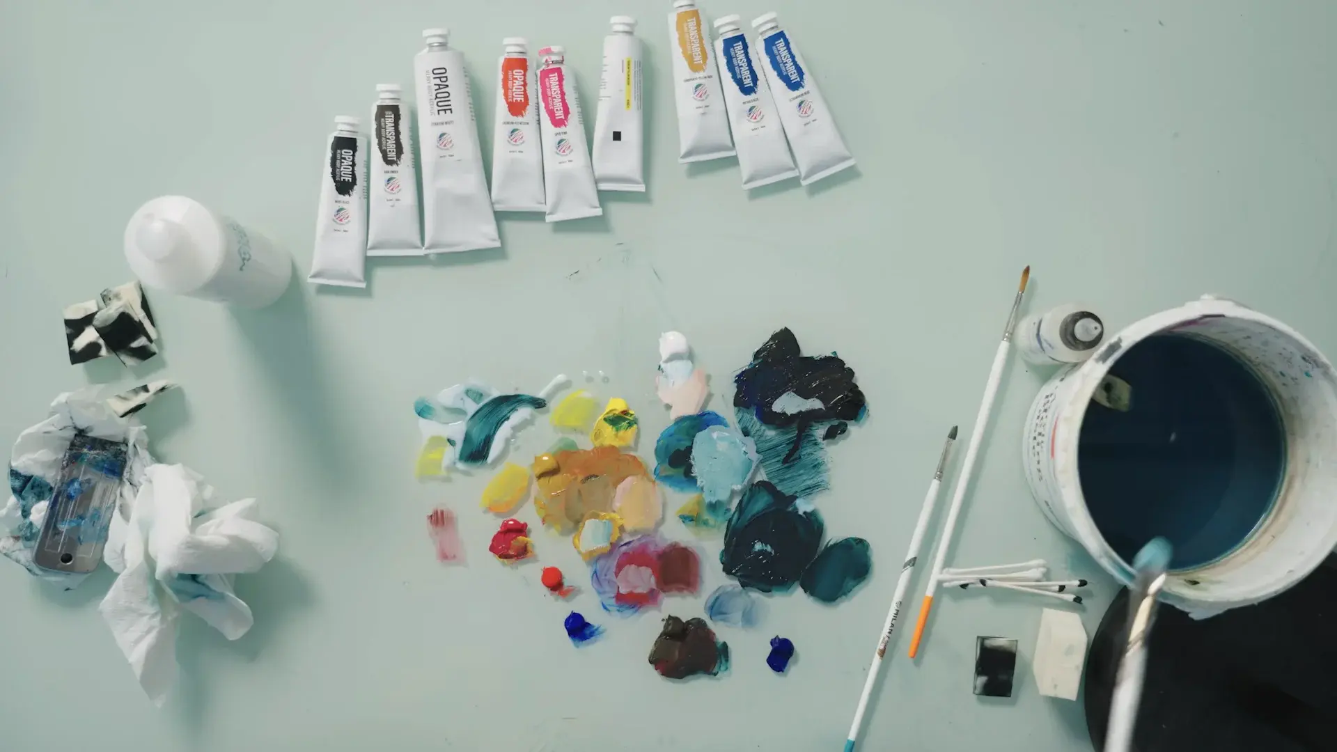 Overhead view of a table with paint tubes, a palette of mixed blues and yellows, brushes and a water bucket